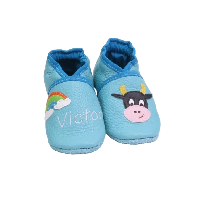 Chaussons en cuir souple artisanaux bleu clair pour bébé avec prénom brodé et motif vache noire et blanche.