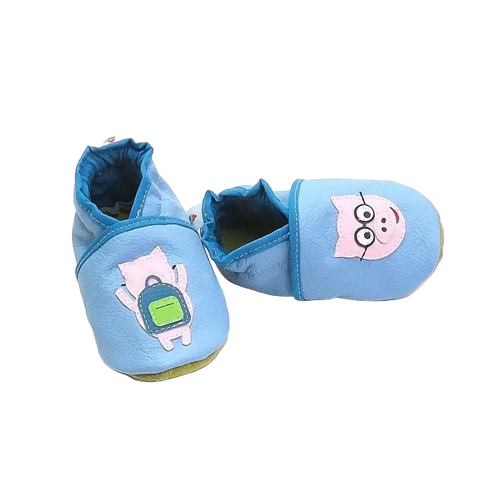Chaussons en cuir souple artisanaux bleu pour bébé avec cochon rose à lunettes sur un pied et cochon avec sac à dos vert sur l’autre, finitions bleu foncé.