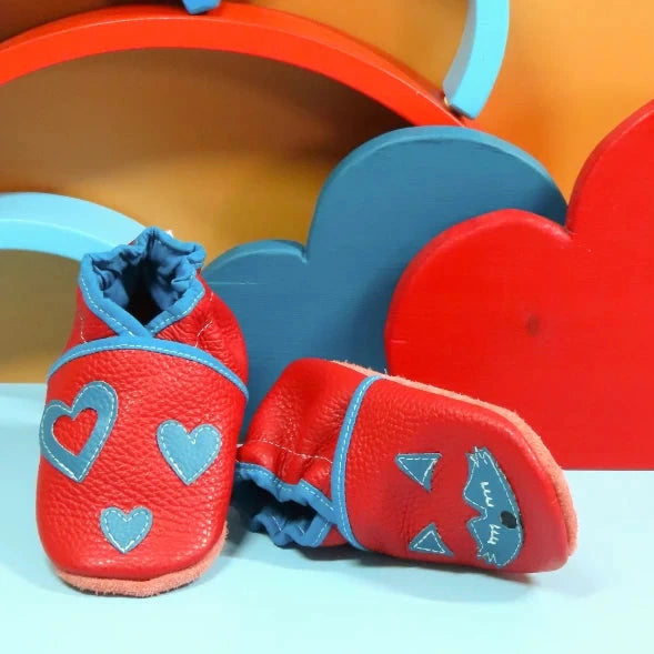 Chaussons en cuir souple artisanaux rouges pour bébé avec renard bleu sur un pied et cœurs bleus sur l’autre.