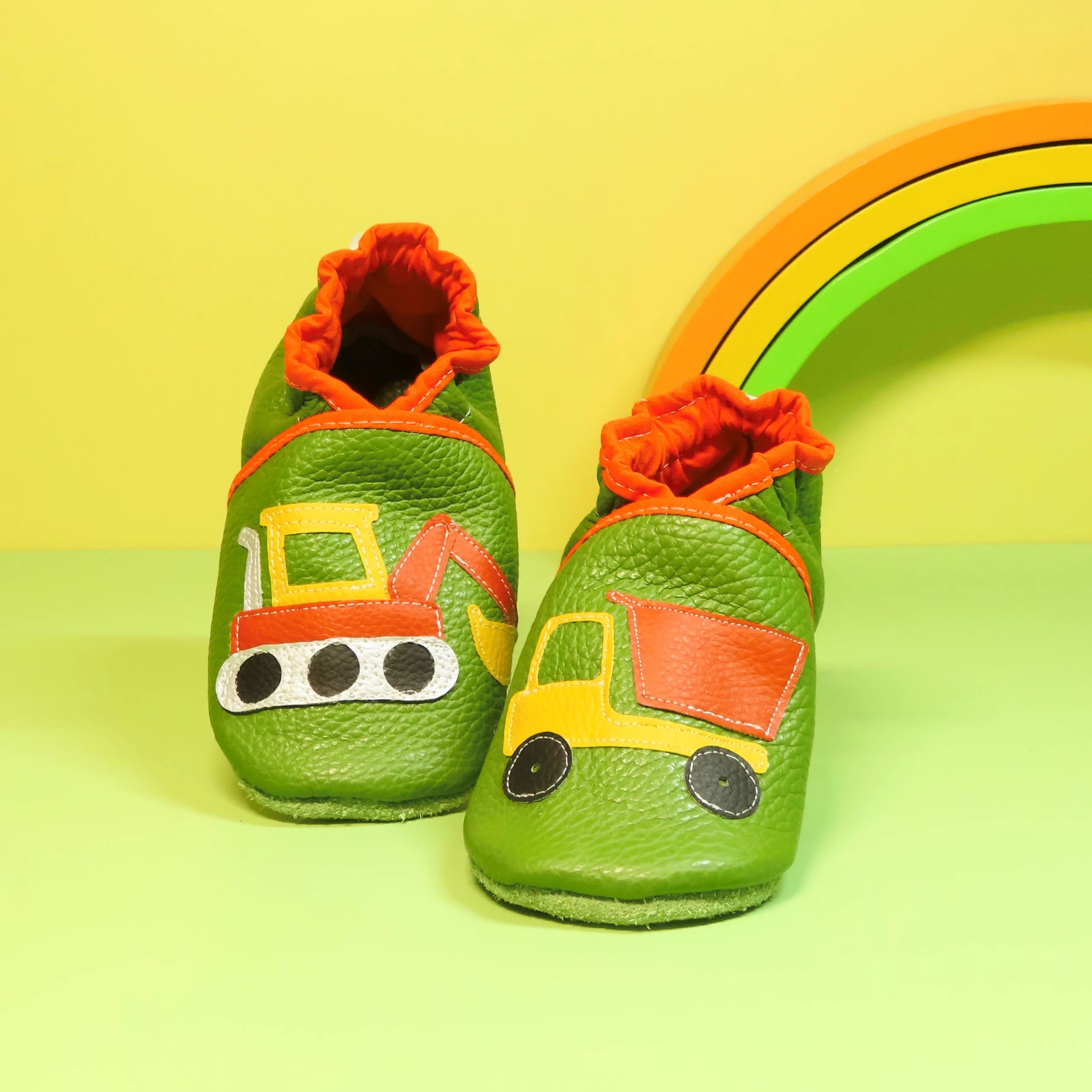 Chaussons en cuir souple artisanaux verts pour enfant, avec pelleteuse sur un pied et camion benne sur l’autre, finitions orange.