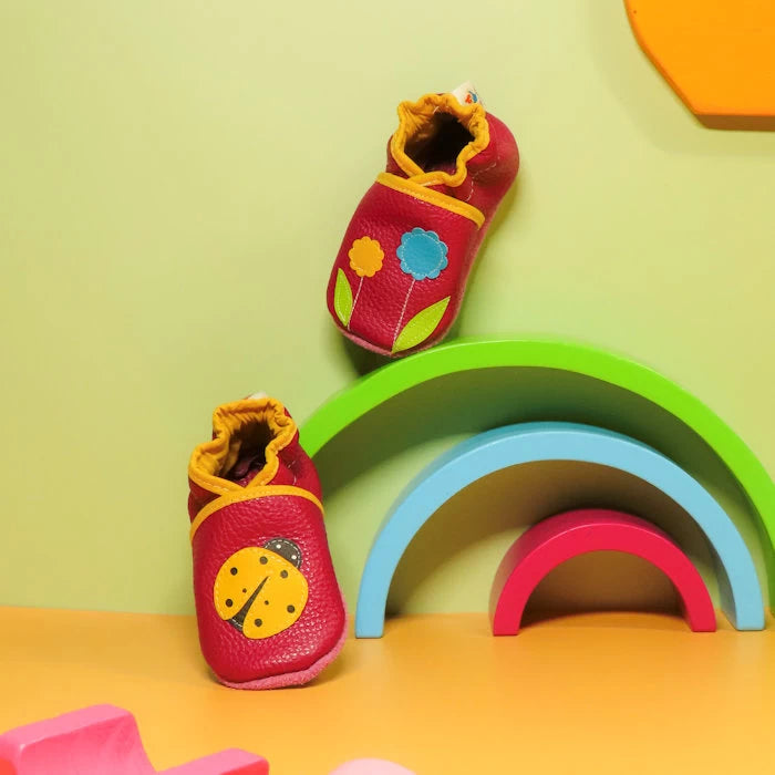 Chaussons bébé en cuir souple rose motif coccinelle et fleurs colorées, mise en scène avec décor arc-en-ciel pastel.