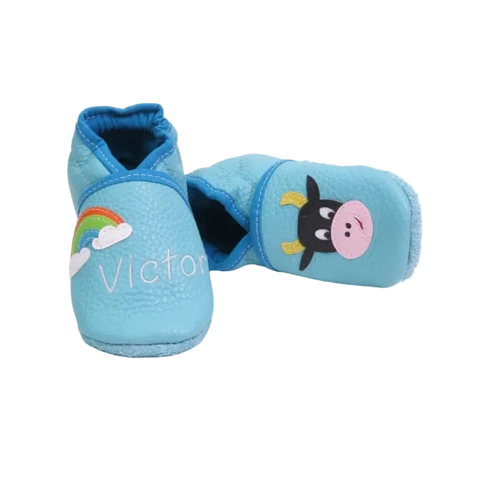 Chaussons en cuir souple artisanaux bleu clair pour bébé avec prénom brodé et motif vache noire et blanche.