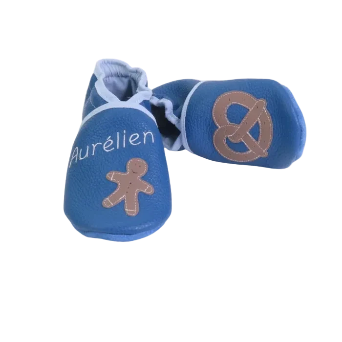 Chaussons en cuir souple artisanaux bleus pour bébé avec prénom personnalisé, petit bonhomme pain d’épice sur un pied et bretzel sur l’autre.