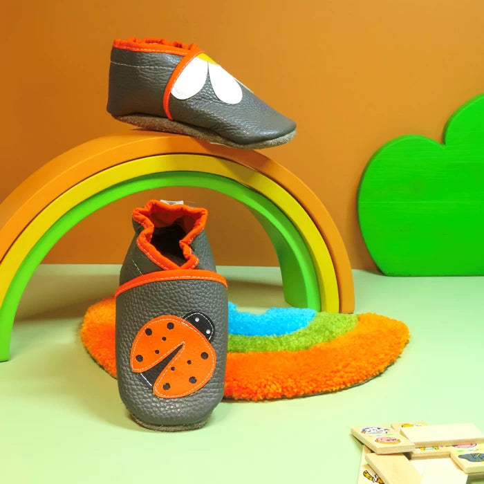 Chaussons bébé en cuir souple gris avec coccinelle orange et pâquerette blanche, présentés sur décor arc-en-ciel.
