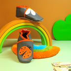 Chaussons bébé en cuir souple gris avec coccinelle orange et pâquerette blanche, présentés sur décor arc-en-ciel.