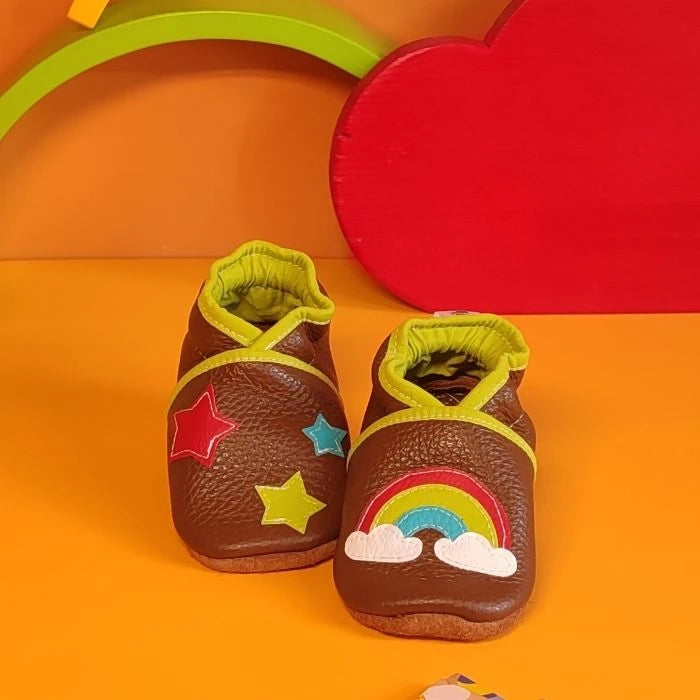 Chaussons en cuir souple marron pour bébé avec arc-en-ciel, nuages et étoiles décoratives, vue croisée mettant en valeur la texture du cuir.