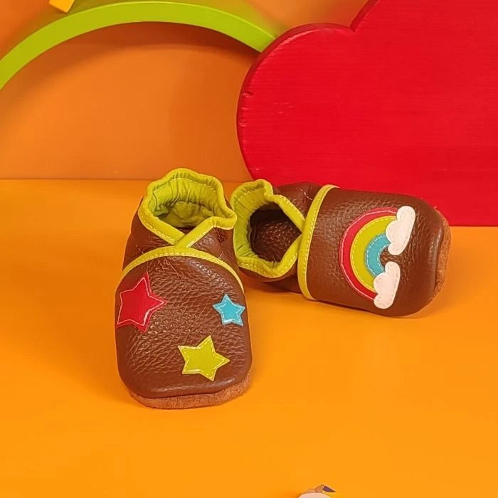 Chaussons en cuir souple marron pour bébé avec arc-en-ciel, nuages et étoiles décoratives, vue croisée mettant en valeur la texture du cuir.
