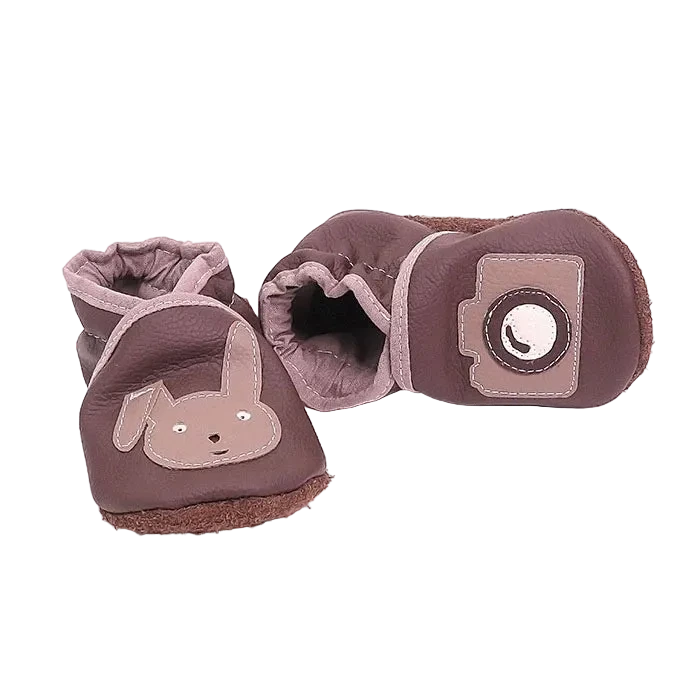 Chaussons en cuir souple artisanaux marron pour bébé avec tête de lapin sur un pied et appareil photo illustré sur l’autre, tons vieux rose.