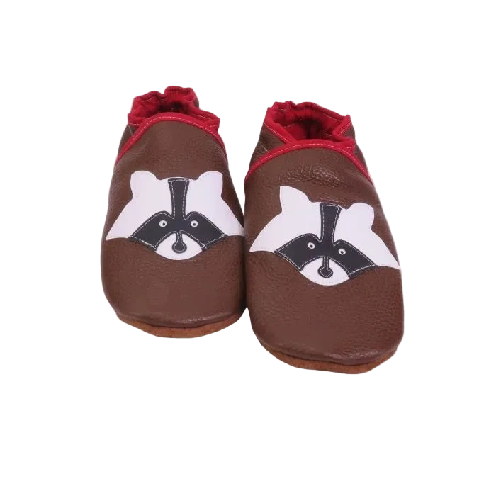 Chaussons en cuir souple artisanaux marron pour bébé avec tête de raton laveur noire et blanche, finitions rouges.