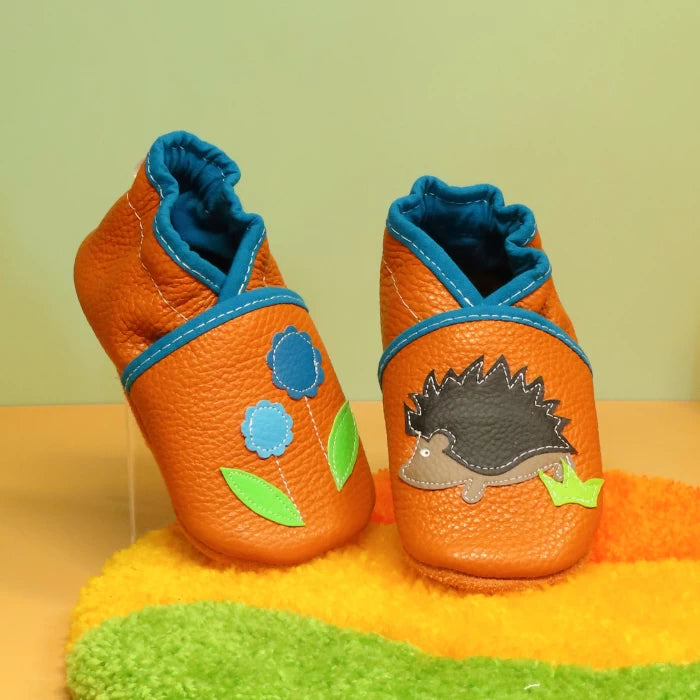 Chaussons bébé en cuir souple orange et bleu avec motif hérisson et fleurs, modèle artisanal coloré.