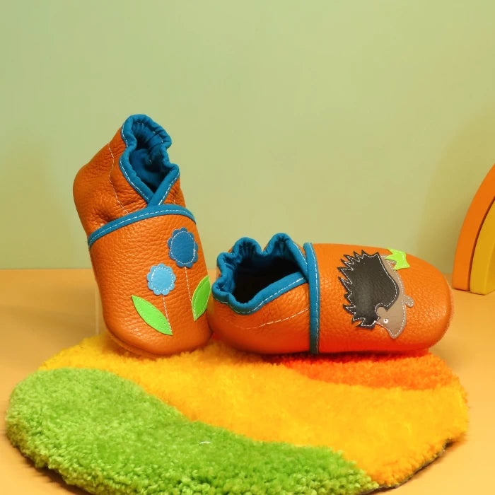 Chaussons en cuir souple pour bébé orange et bleu avec hérisson appliqué, vue de profil mettant en valeur la souplesse du cuir.