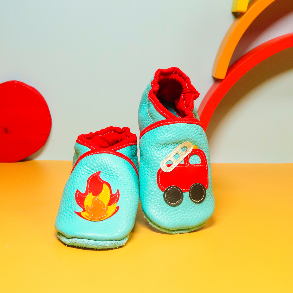 Chaussons bébé en cuir souple bleu menthe avec camion de pompier rouge et flamme appliquée, photographiés en mise en scène colorée.
