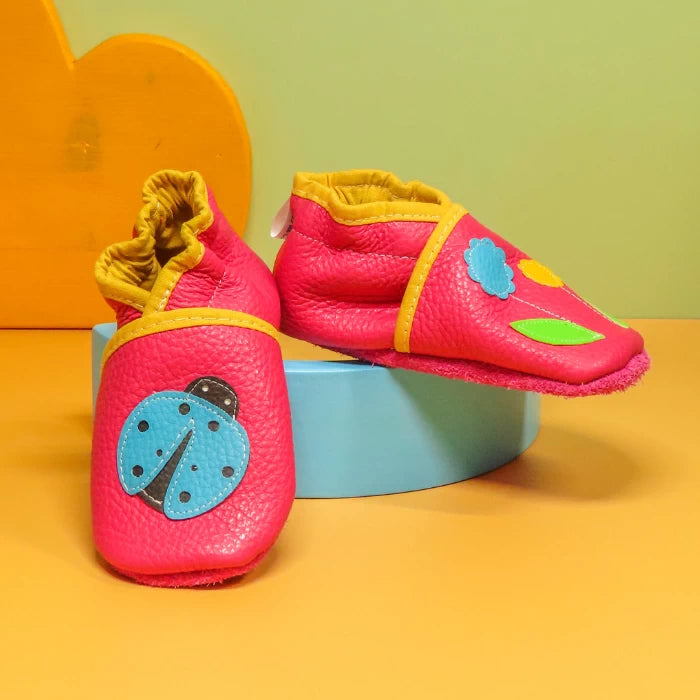 Chaussons en cuir souple artisanaux rose pour bébé avec coccinelle bleue sur un pied et fleurs colorées sur l’autre, vue en biais sur socle bleu.