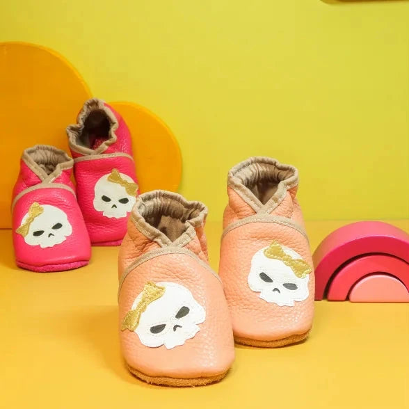 Chaussons bébé tête de mort avec nœud doré et rose, photo d’ambiance.