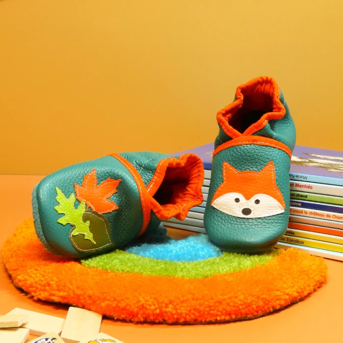 Chaussons bébé en cuir souple vert d’eau avec motif renard et feuilles d’automne, présentés sur des livres colorés.