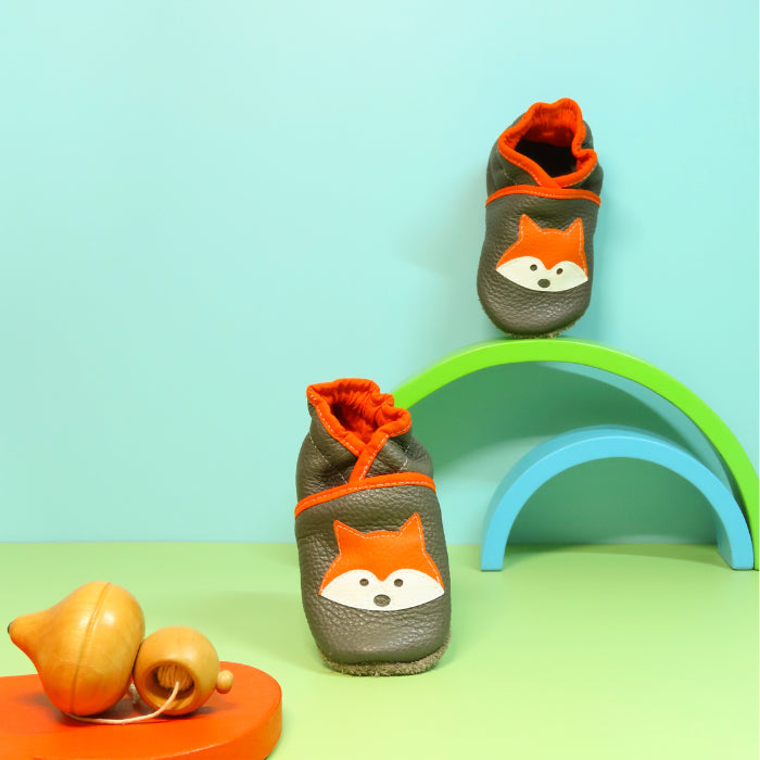 Chaussons en cuir souple artisanaux gris pour enfant avec motif renard orange, mise en scène colorée avec arche verte.