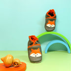 Chaussons en cuir souple artisanaux gris pour enfant avec motif renard orange, mise en scène colorée avec arche verte.