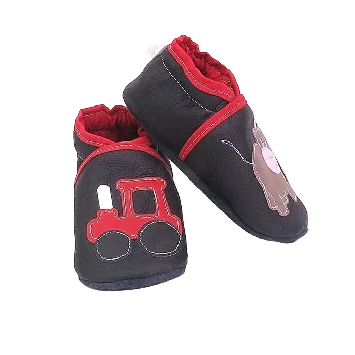 Chaussons en cuir souple artisanaux noirs pour enfant, vue en angle, avec tracteur rouge sur un pied et petit taureau marron sur l’autre, finitions rouges.