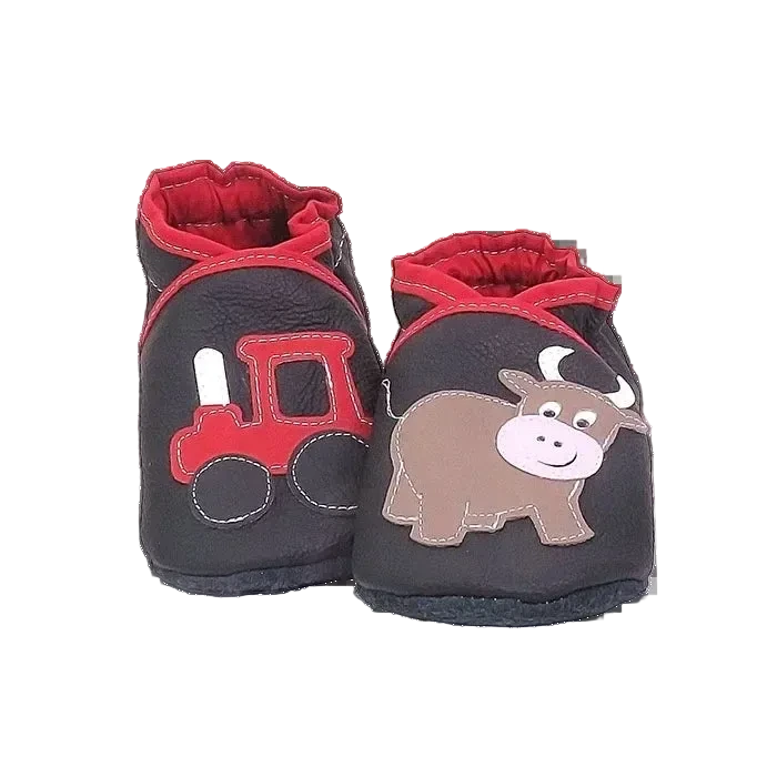 Chaussons en cuir souple artisanaux noirs pour enfant avec tracteur rouge sur un pied et petit taureau marron sur l’autre, finition élastiquée rouge.