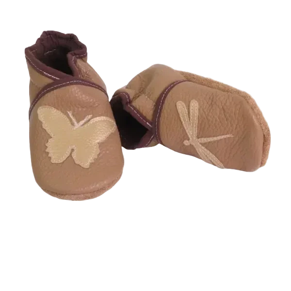 Chaussons en cuir souple artisanaux couleur camel pour enfant, vue en angle, avec papillon beige sur un pied et libellule beige sur l’autre, finitions marron.