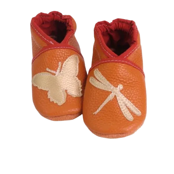Chaussons en cuir souple artisanaux orange pour enfant, avec papillon crème sur un pied et libellule crème sur l’autre, finition élastiquée .