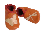Chaussons en cuir souple artisanaux orange pour enfant, vue en angle, avec papillon crème sur un pied et libellule crème sur l’autre, finitions rouges élastiquées.