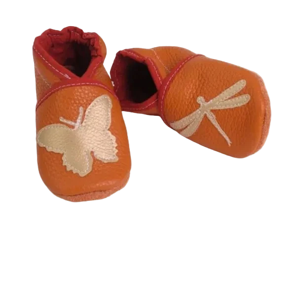 Chaussons en cuir souple artisanaux orange pour enfant, vue en angle, avec papillon crème sur un pied et libellule crème sur l’autre, finitions rouges élastiquées.