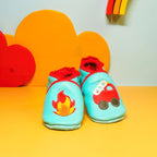 Chaussons bébé en cuir souple bleu menthe avec camion de pompier rouge et flamme appliquée, photographiés en mise en scène colorée.